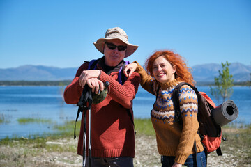 A man and a woman are standing by a lake, smiling and posing for a picture. They are hiking or trekking.