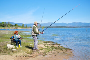 A man and a woman are fishing in a lake. The man is holding a fishing rod and the woman is sitting in a chair eating a sandwich. Scene is peaceful and relaxing