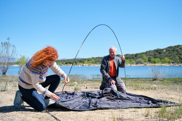 A man and a woman are setting up a tent by a lake. They are working as a team