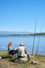 A man and a woman are sitting by a lake, fishing. The man is looking at the fishing rod and the woman is reading a book. The scene is peaceful and relaxing