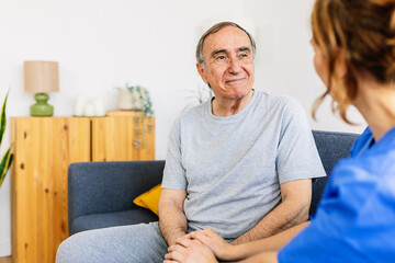Female caregiver talking with senior male patient at nursing home. Young doctor holding hands with old man on sofa, supporting, sitting together on sofa. Elderly healthcare and assistance concept.