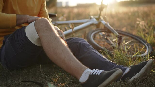 Unrecognizable man wraps bandage around knee after receiving injury. Cyclist administers first aid to himself after falling and injuring knee in rural environment. Fixing joint with tight bandage.