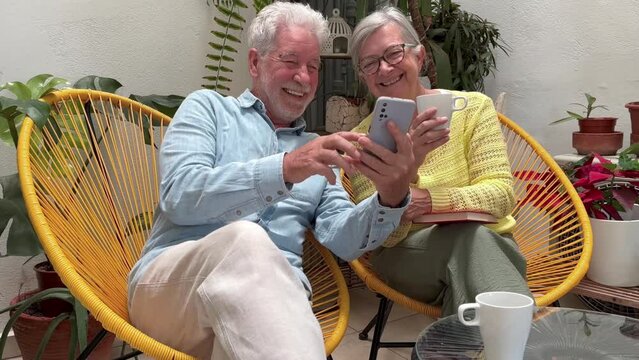 Senior adult couple sitting on home balcony relaxing with a coffee cup, retired seniors looking together at the same mobile phone smiling share good of fun news