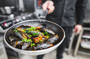 Close-up of a chef’s hands holding a pan with cooked mussels garnished with herbs, suggesting a fresh seafood dish in a kitchen setting