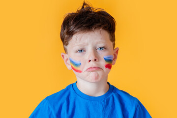 Young boy sad with colorful face paint on yellow background