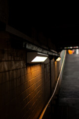 A dark subway with two street lights shining on the brick wall. The light is yellow and the wall is made of bricks