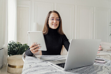 A professional woman with glasses, wearing a black sweater, multitasking with a laptop and tablet.
