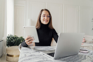 Fototapeta premium A professional woman with glasses, wearing a black sweater, multitasking with a laptop and tablet.
