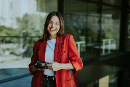 Smiling young business woman in red blazer enjoying morning coffee outdoors in modern urban setting