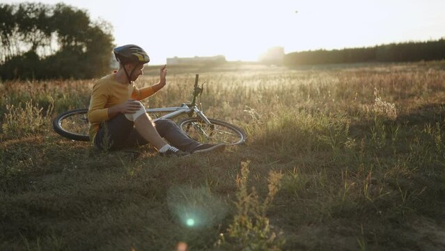 Push in shot of frustrated cyclist after falling and getting injured. Man in helmet annoyedly hits bicycle with hand and rubs knee while sitting in field at sunset. Sports risks.