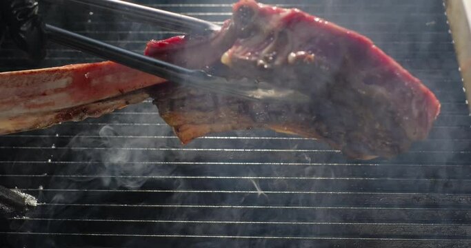 Juicy tomahawk steak grilling on barbecue grid. Chef hand flips roasting steak. Grilling BBQ meat