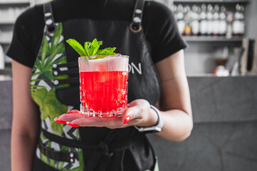 Close-up of a hand holding a vibrant red cocktail with fresh mint garnish, against a blurred bar...