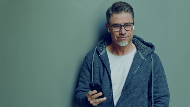 A mature man in casual hooded sweater focusing on cell phone, hearing music in earbuds, leaning against a plain gray wall. Portrait of mid adult male in 50s, bearded, grey hair.