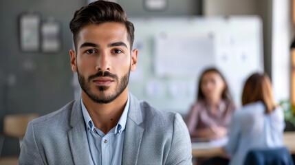 Obraz premium A man with a beard and short hair wearing a light blue shirt and a gray blazer sitting in an office environment with blurred colleagues in the background.