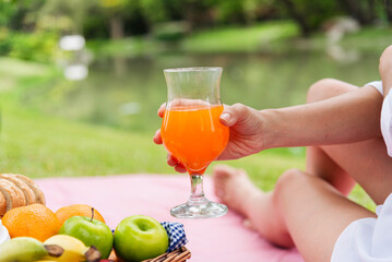 Close up Women picnic in green park hands holding glass of orange juice fresh fruit cool summer. Women drinking sit blanket with Picnic basket bread coffee cup in wood wicker healthy box red Blanket