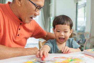 Asian Senior man and Grandson or young boy laughing and drawing together at home, showcasing...