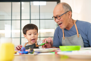 Asian Senior man and young child boy or Grandson enjoying painting together at a table, emphasizing...