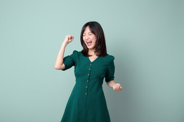 Young overjoyed happy excited woman of Asian ethnicity 30s wear green dress do winner gesture celebrate clenching fists say yes isolated on pastel plain green color background studio portrait.