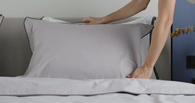 Woman hand set up pillow on bed sheet in bedroom. Young female hands gently touch grey pillow