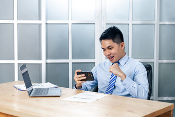 Asian businessman working in his office using a mobile smart phone feeling happy and excited while fist up cheering in success, professional business worker with a computer laptop sitting at his desk.