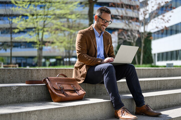 Full length of male entrepreneur reading e-mails over laptop while sitting with bag on steps in city