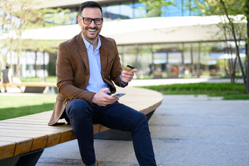 Portrait of smiling handsome employee with credit card and mobile phone sitting on bench in city