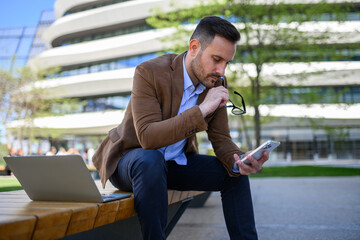 Young businessman holding glasses and reading messages over phone while sitting with laptop on bench