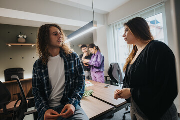 A young man and woman engage in a business discussion at an office table, with coworkers collaborating in the background.