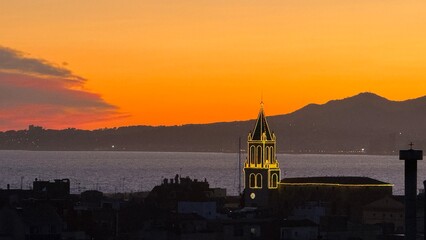 Illuminated church bell tower against an orange coastal sunset and mountain silhouette © ZEDRAY