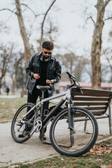Fototapeta premium A fashionable male business entrepreneur pausing to use his smart phone while standing by his bike in a scenic park setting.
