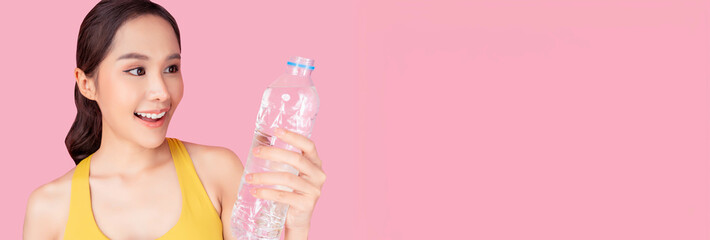 A smiling young woman in a yellow sports bra holding a bottle of water against a pink background....