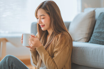 Joyful young asian female enjoying a cup of coffee while sitting on the rug beside to the sofa at home, Cosy scene, Smiling pretty woman drinking hot tea