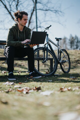 A good-looking male enjoys a relaxing day working outdoors on his laptop, seated beside his bicycle in a sunny park.