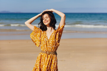 Elegant woman in a sunny yellow dress standing on the sandy beach with hands on head