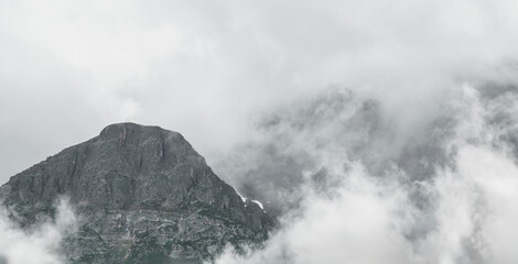 Dramatic capture of a large gray rocky mountain that is surrounded by white to gray clouds and mist.  There is ample room for copy,

