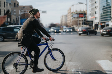 A young man wearing a beanie pedals a blue bicycle across an urban street with cars and city buildings in the background.