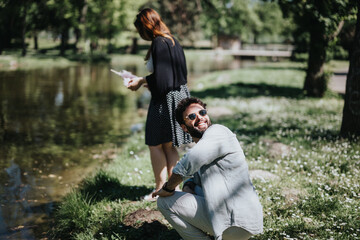 A happy man on one knee proposing to a surprised woman in a park with a ring on a sunny day,...