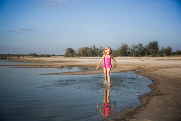 A little seven-year-old girl in a pink swimsuit runs along the lake in a good cheerful mood with a...