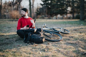 A cheerful young man in a red shirt relaxes in a grassy park, holding a smart phone with a mountain bike lying beside him.