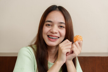 Asian teenage girl with long brown hair holding peeled orange, showing braces, smiling brightly. Wearing light green sweater, sitting indoors. cheerful and positive demeanor.