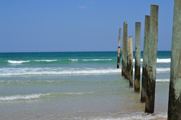 Waves break upon a series of wooden pole pilings, set against the backdrop of a turquoise sea and...