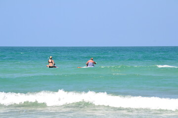 Two women are paddleboarding in the turquoise sea, with waves breaking along the water's edge under...