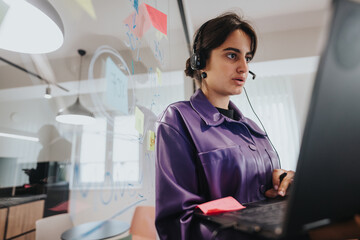 An attentive young woman wearing a headset is engaged with a computer in a vibrant office setting,...