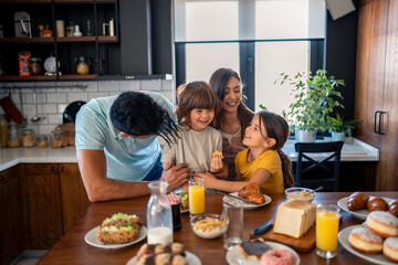 Happy parents having breakfast at kitchen table with children at home, looking at them with love...