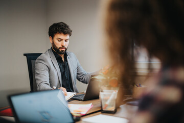 A young, focused man with a beard working at his laptop during a business meeting in a modern...