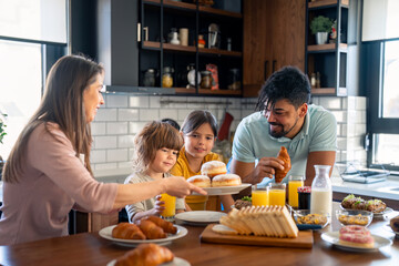 Kids at breakfast with parents. Mother is serving donuts on a plate. Cheerful family moments at home.