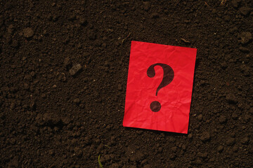 A red paper note with a question mark on it lying on a background of soil. Close up.