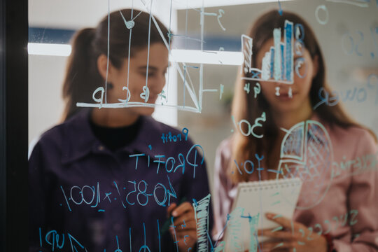 Two businesswomen engage in a strategic session, interpreting and discussing complex statistics written on a transparent glass wall using markers and sticky notes.