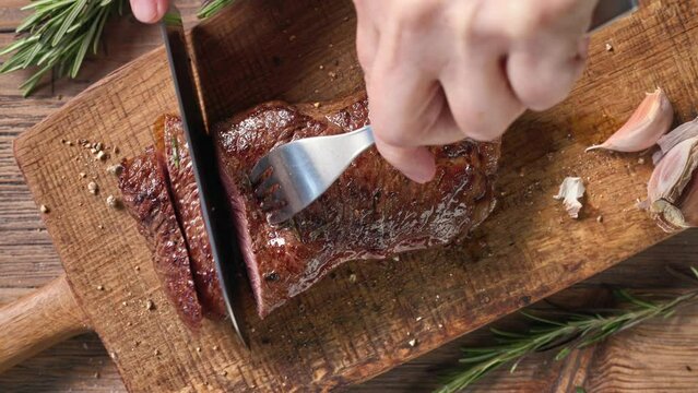 Chef slicing freshly grilled juicy beef steak meat with a knife on a wooden cutting board in the kitchen, food close-up