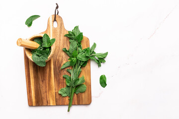Wooden mortar with fresh green mint leaves on the wooden board on white marble table top view, copy space for your design. Herbal and aromatheraphy concept.
